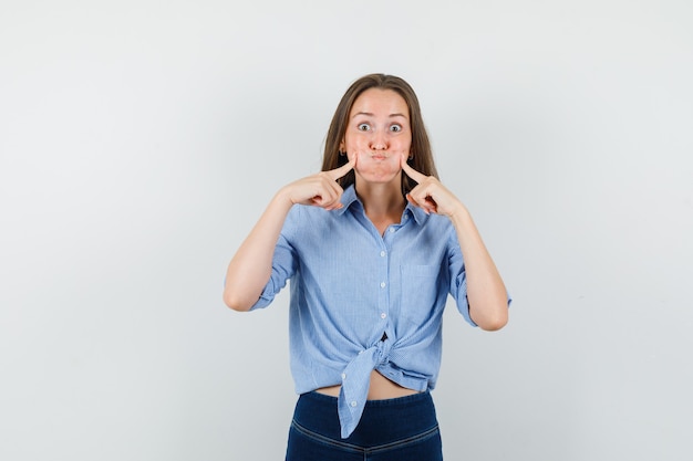 Giovane donna in camicia blu, pantaloni premendo sulle guance che soffia e guardando divertente