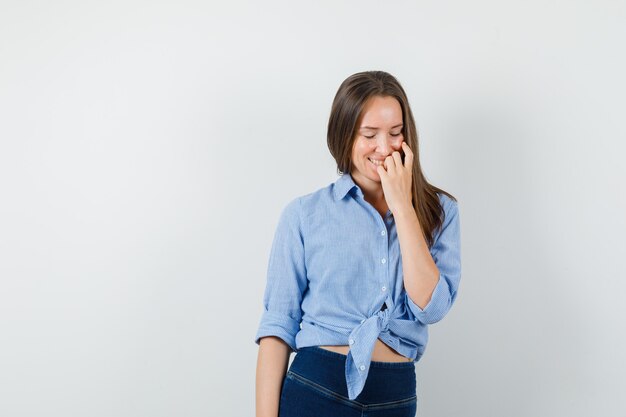 Giovane donna in camicia blu, pantaloni tenendo la mano sul viso e guardando gioiosa