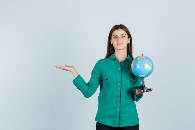 Giovane donna in camicia che tiene il globo terrestre, facendo il gesto di benvenuto e guardando soddisfatto, vista frontale.