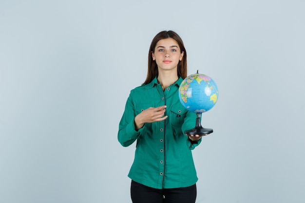 Giovane donna in camicia che tiene il globo terrestre, indicandolo e guardando fiducioso, vista frontale.
