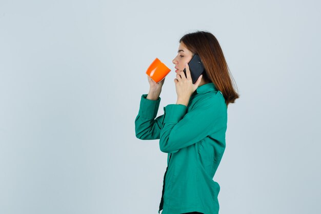 Giovane donna in camicia che tiene tazza di tè arancione, parlando al telefono cellulare e guardando fiducioso, vista frontale.