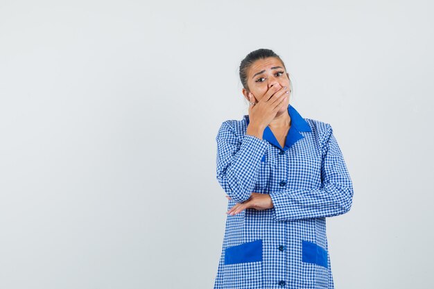 Giovane donna in camicia del pigiama a quadretti blu che copre la bocca con le mani e guardando sorpreso, vista frontale.
