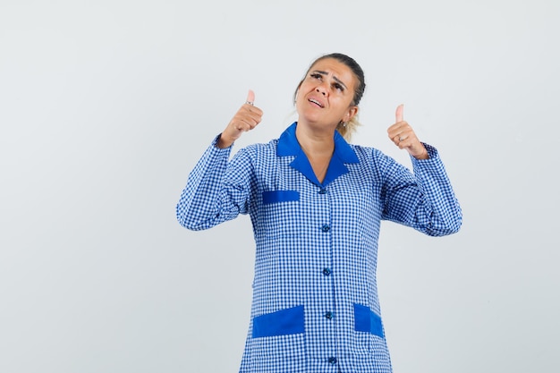 Giovane donna in camicia del pigiama a quadretti blu che mostra i pollici in su con entrambe le mani e sembra carina, vista frontale.