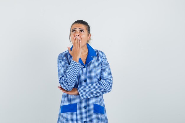 Giovane donna in camicia del pigiama a quadretti blu mettendo la mano sulla bocca, in piedi nel gesto di pensiero e guardando pensieroso, vista frontale.