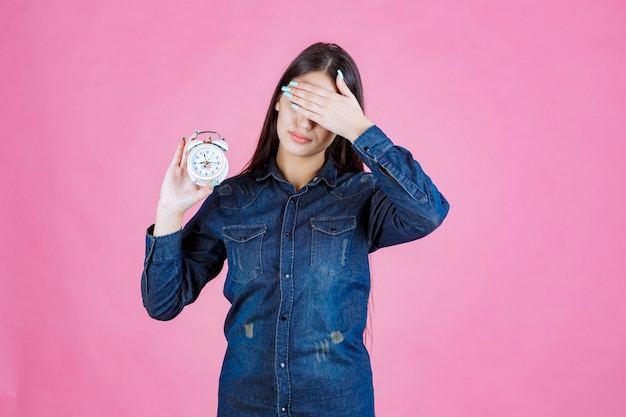 Giovane donna in camicia di jeans che tiene la sveglia e che copre gli occhi