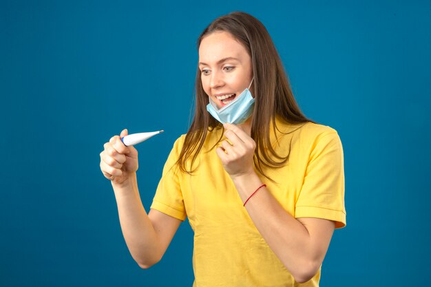 Giovane donna in camicia di polo gialla che toglie maschera protettiva medica che sorride esaminando termometro con il fronte felice isolato su fondo blu