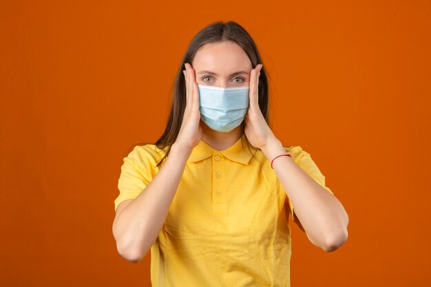 Giovane donna in camicia di polo gialla e maschera protettiva medica toccando il viso guardando la telecamera su sfondo arancione