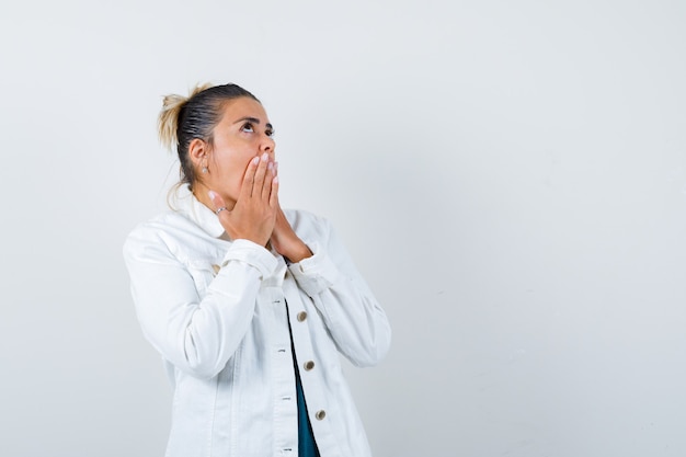 Giovane donna in camicia, giacca bianca con le mani sulla bocca e sguardo pensieroso, vista frontale.