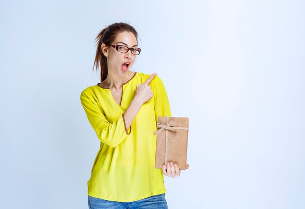 Giovane donna in camicia gialla che tiene in mano una scatola regalo di cartone e indica qualcuno sulla destra