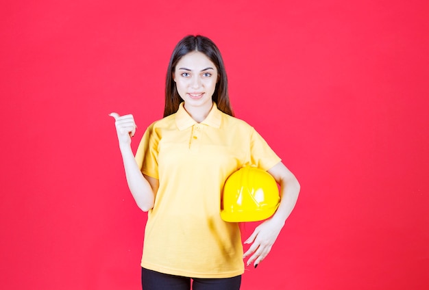 Giovane donna in camicia gialla che tiene un casco giallo e indica qualcuno dietro