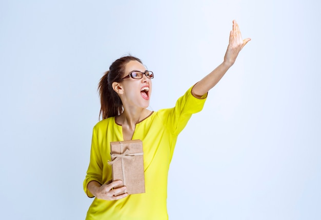 Giovane donna in camicia gialla che tiene una scatola regalo di cartone e invita una persona a presentarla