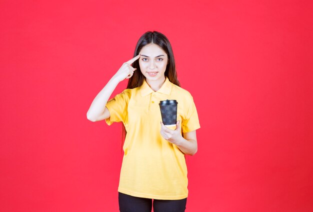 Giovane donna in camicia gialla che tiene una tazza di caffè usa e getta nera, pensando e avendo una buona idea