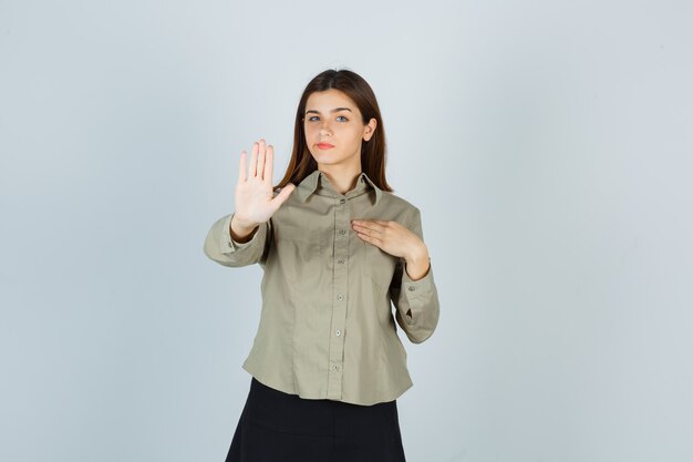 Giovane donna in camicia, gonna che mostra il gesto di rifiuto, tenendo la mano sul petto