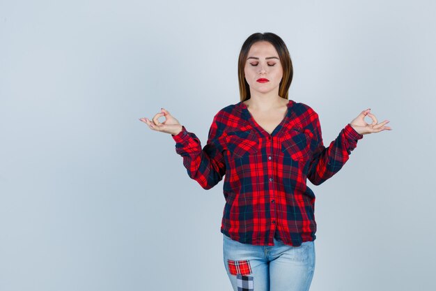 Giovane donna in camicia, jeans che mostra il segno mudra mentre tiene gli occhi chiusi e sembra rilassata, vista frontale.