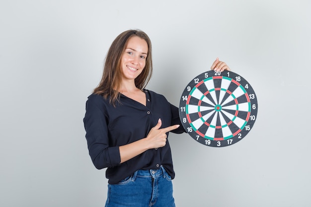 Giovane donna in camicia nera, pantaloncini di jeans che punta il dito al bersaglio e sembra allegra