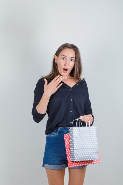 Giovane donna in camicia nera, pantaloncini di jeans che tengono i sacchetti di carta e che sembrano sorpresi