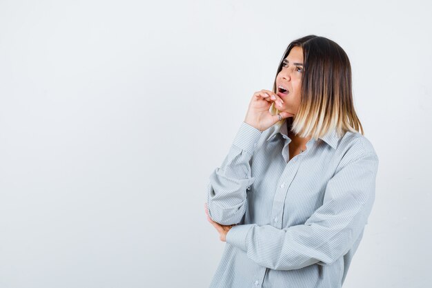 Giovane donna in camicia oversize che tiene la mano sotto il mento e sembra perplessa, vista frontale.