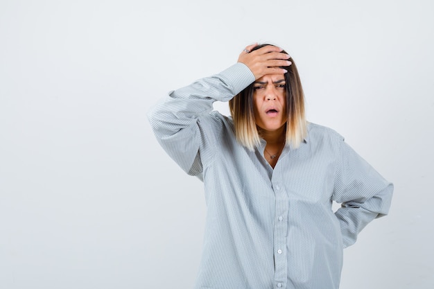 Giovane donna in camicia oversize che tiene la mano sulla fronte mentre tiene la mano sull'anca e sembra smemorata, vista frontale.