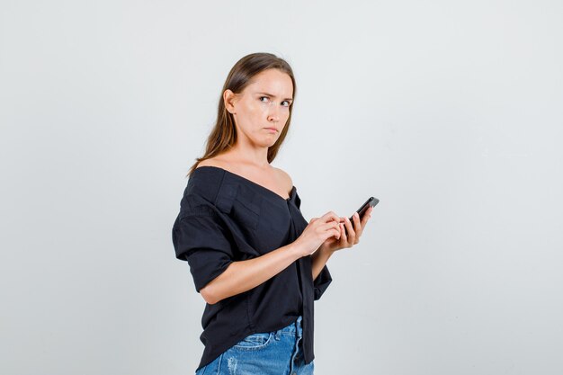 Giovane donna in camicia, pantaloncini alla ricerca da parte mentre si tiene lo smartphone e sembra seria