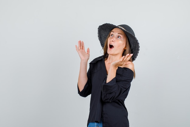 Giovane donna in camicia, pantaloncini, cappello alzando le mani e guardando sorpreso