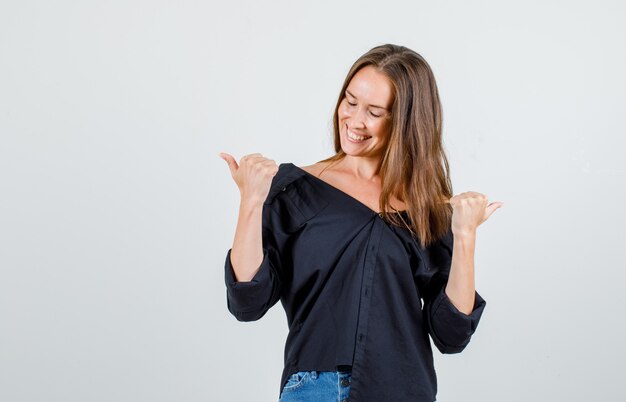 Giovane donna in camicia, pantaloncini che punta i pollici di distanza e sembra allegra