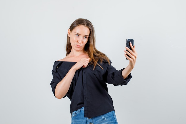 Giovane donna in camicia, pantaloncini in posa mentre prende selfie sullo smartphone e sembra carino