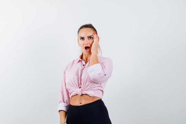 Giovane donna in camicia, pantaloni tenendo la mano sulla guancia e guardando stupito