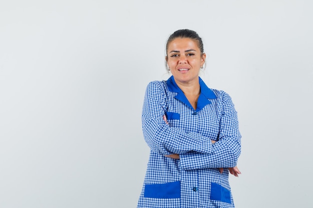 Giovane donna in camicia pigiama a quadretti blu in piedi con le braccia incrociate e sorridente e guardando felice, vista frontale.