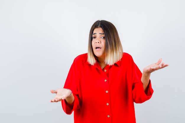 Giovane donna in camicia rossa oversize che mostra gesto impotente e sembra ansiosa, vista frontale.