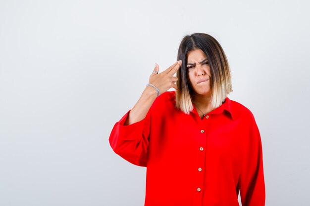 Giovane donna in camicia rossa oversize che mostra il gesto della pistola e sembra seria, vista frontale.