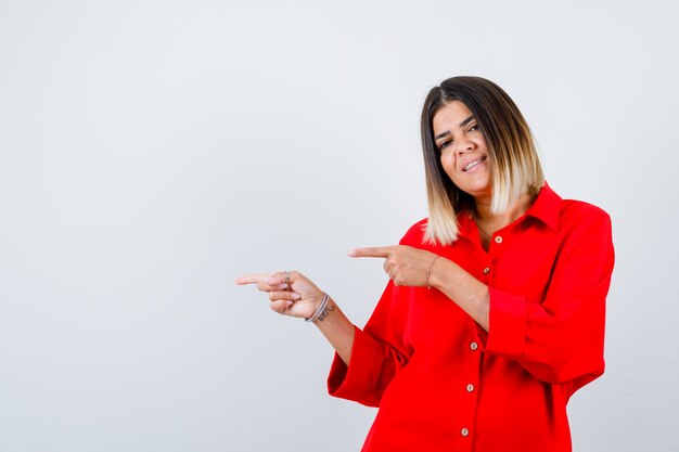 Giovane donna in camicia rossa oversize che punta al lato sinistro e sembra soddisfatta, vista frontale.