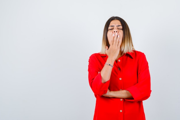 Giovane donna in camicia rossa oversize che sbadiglia e sembra assonnata, vista frontale.