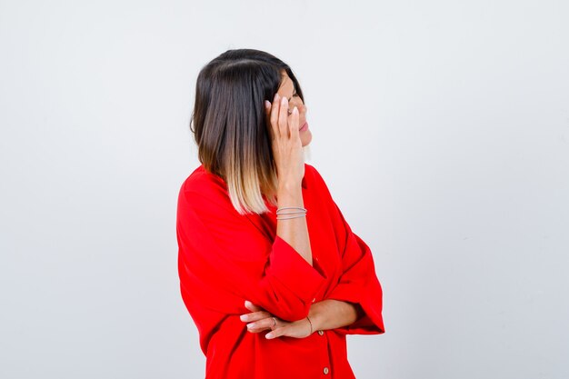 Giovane donna in camicia rossa oversize che si appoggia la testa a portata di mano e sembra stanca, vista frontale.