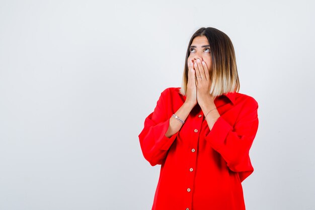 Giovane donna in camicia rossa oversize con le mani sulla bocca e con sguardo concentrato, vista frontale.