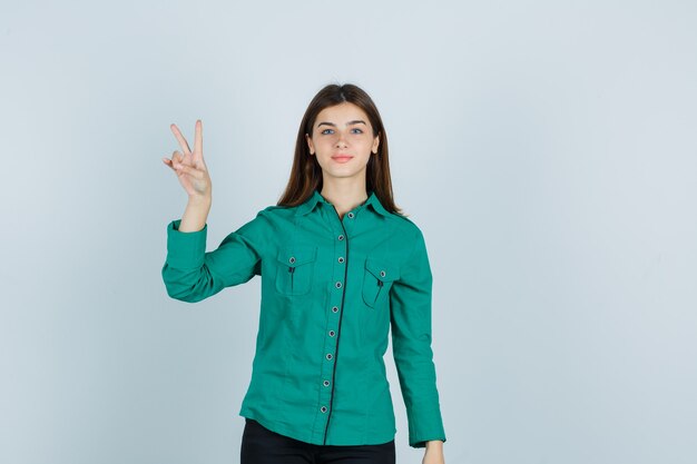 Giovane donna in camicia verde che mostra il gesto di vittoria e guardando fiducioso, vista frontale.