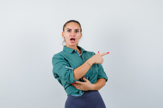 Giovane donna in camicia verde che punta a destra e sembra scioccata, vista frontale.