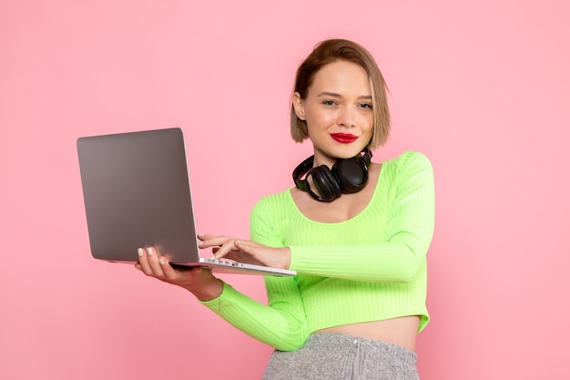 giovane donna in camicia verde e pantaloni grigi in posa con laptop e auricolari neri