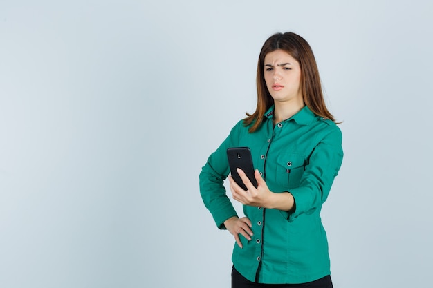 Giovane donna in camicia verde guardando il telefono cellulare e guardando perplesso, vista frontale.