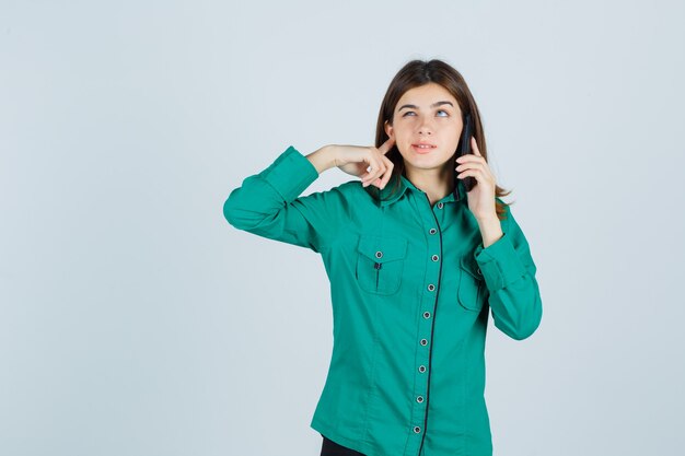 Giovane donna in camicia verde parlando al telefono cellulare, tappando l'orecchio con il dito e guardando confuso, vista frontale.