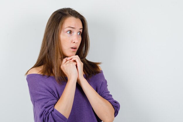 Giovane donna in camicia viola puntellando il mento sulle mani giunte e guardando sorpreso, vista frontale.