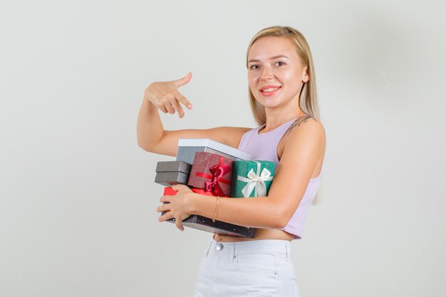 Giovane donna in canottiera, minigonna che punta il dito contro scatole regalo e sembra allegra