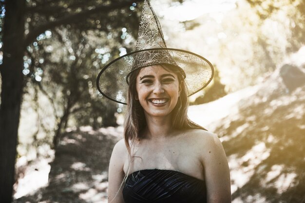 Giovane donna in cappello della strega in piedi nella foresta e sorridente
