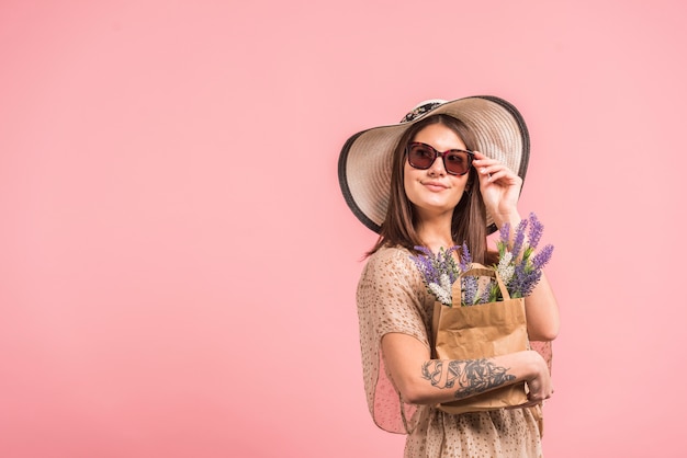 Giovane donna in cappello e occhiali da sole che tiene la borsa con i fiori