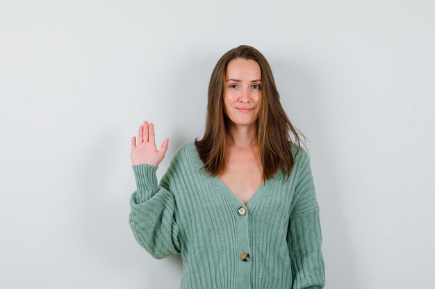 Giovane donna in cardigan di lana agitando la mano per il saluto e guardando fiducioso, vista frontale.