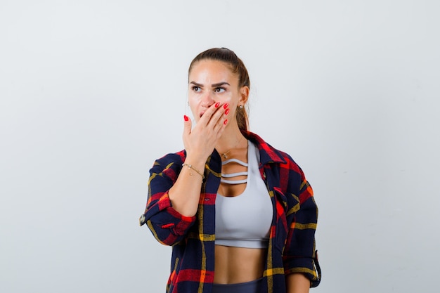 Giovane donna in crop top, camicia a scacchi, pantaloni con la mano sulla bocca e sguardo perplesso, vista frontale.