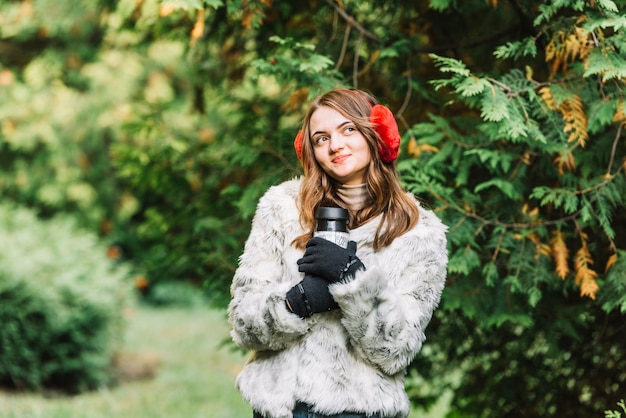 Giovane donna in cuffia che tiene thermos nel parco di conifere