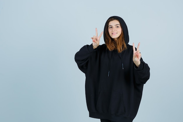 Giovane donna in felpa con cappuccio oversize, pantaloni che mostrano il segno di vittoria e sembra fortunata, vista frontale.