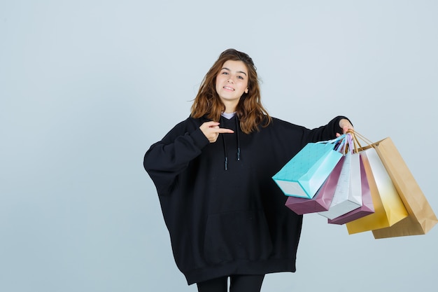 Giovane donna in felpa con cappuccio oversize, pantaloni che punta i pacchetti e che sembra gioiosa, vista frontale.