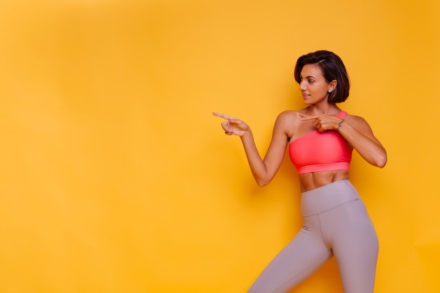 Giovane donna in forma abbastanza forte vestita con abiti sportivi, top alla moda e leggings, posa contro il muro giallo