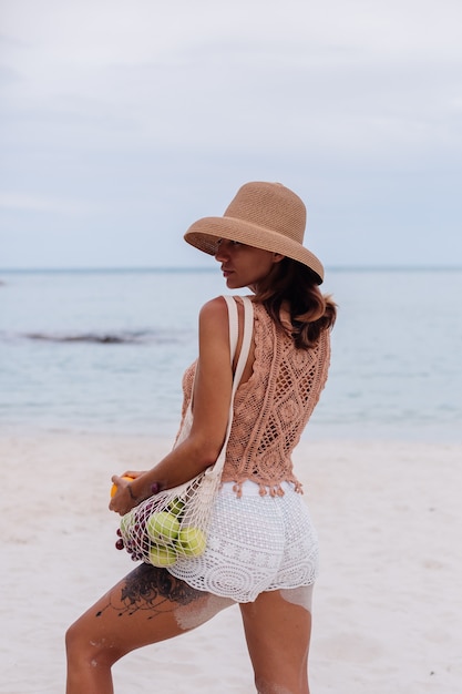 Giovane donna in forma abbronzata abbastanza caucasica in vestiti a maglia e cappello sulla spiaggia